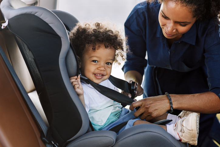 side-view-of-a-happy-little-boy-looking-at-camera-while-his-mother-buckling-him-in-a-car-seat